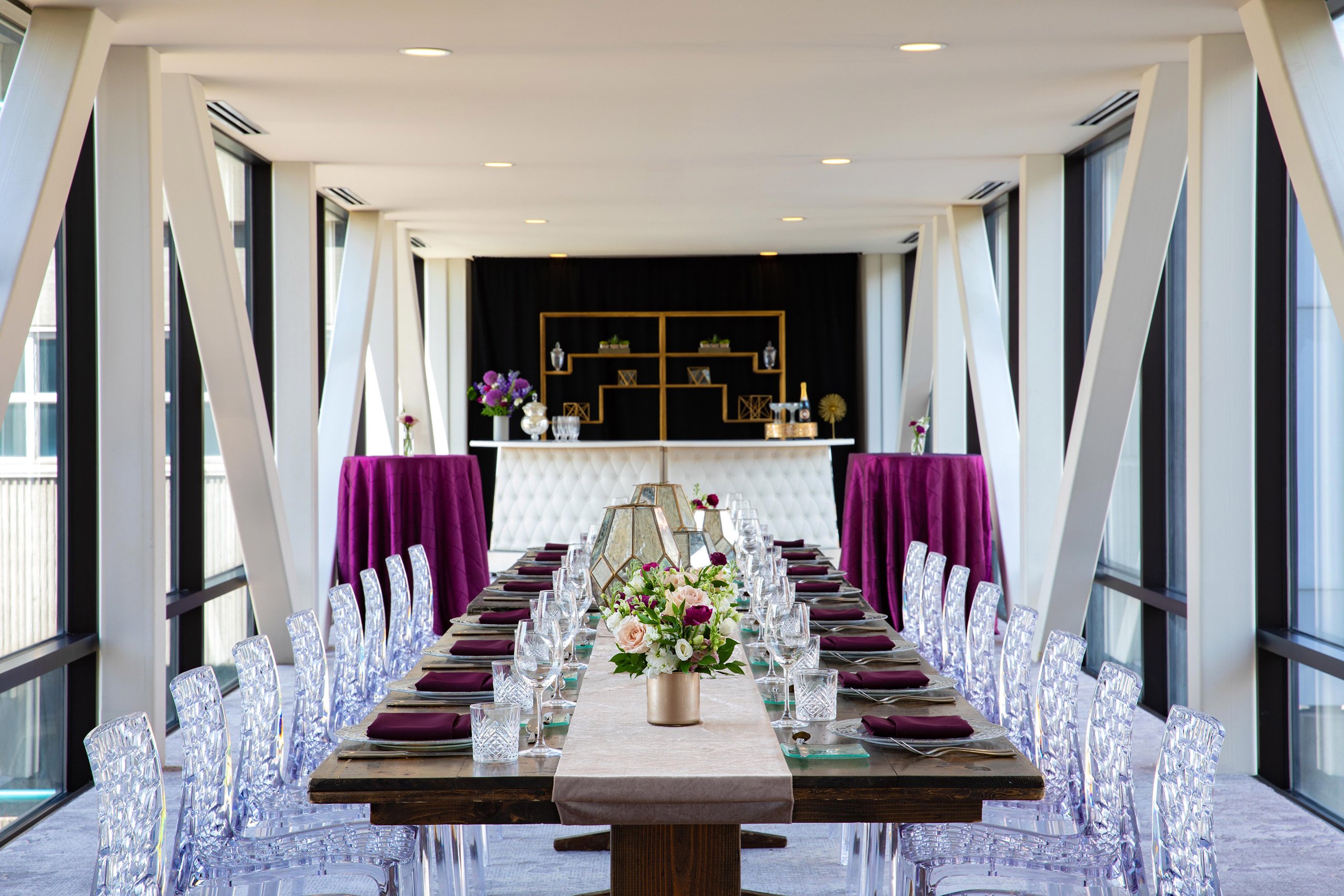 A wooden table with dining settings next to elegant transparent chairs and standing tables with burgundy cloth in Skyway venue at Hotel Ivy