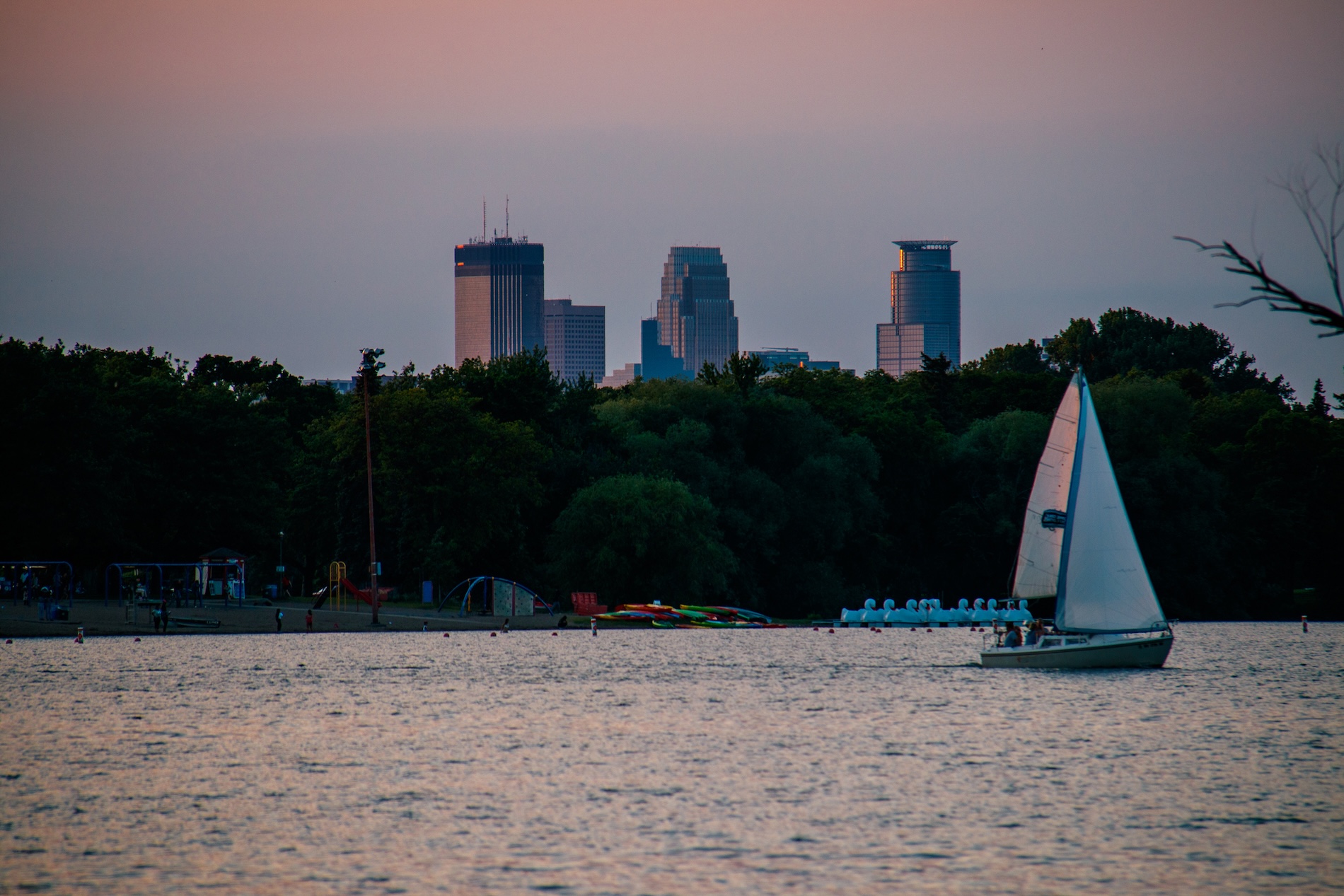 Horizontal color photograph of a sailboat on Lake Nokomis crossing in front of the Minneapolis skyline at sunset.