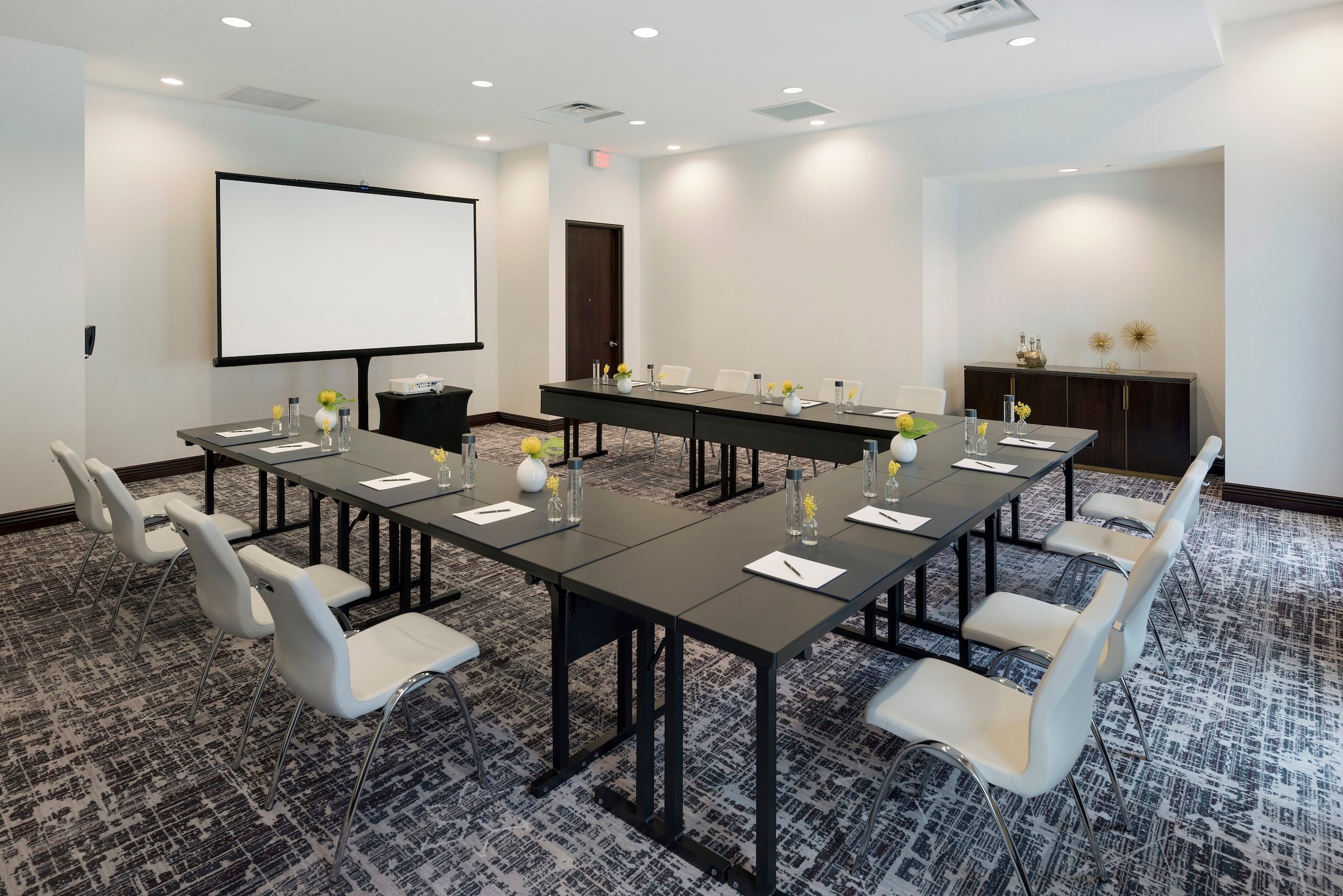 An elegantly designed meeting room at Hotel Ivy’s venue, Exhibit 1 & 2 , set up in a U-shape conference style with white chairs, dark desks, and a presentation screen