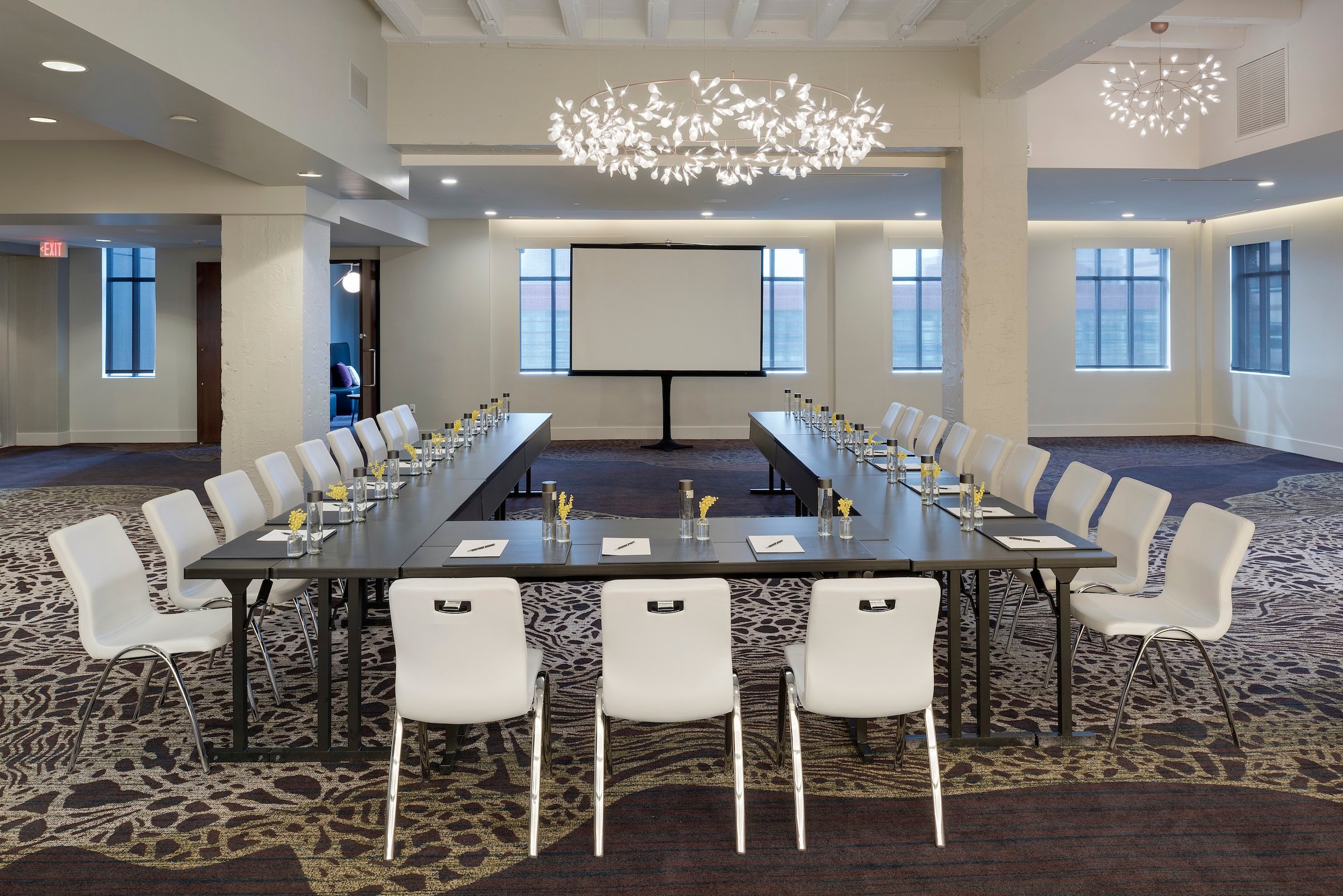 A spacious and elegantly designed meeting room at Hotel Ivy’s Grand Studio, set up in a U-shape conference style with white chairs, dark tables, and a presentation screen.