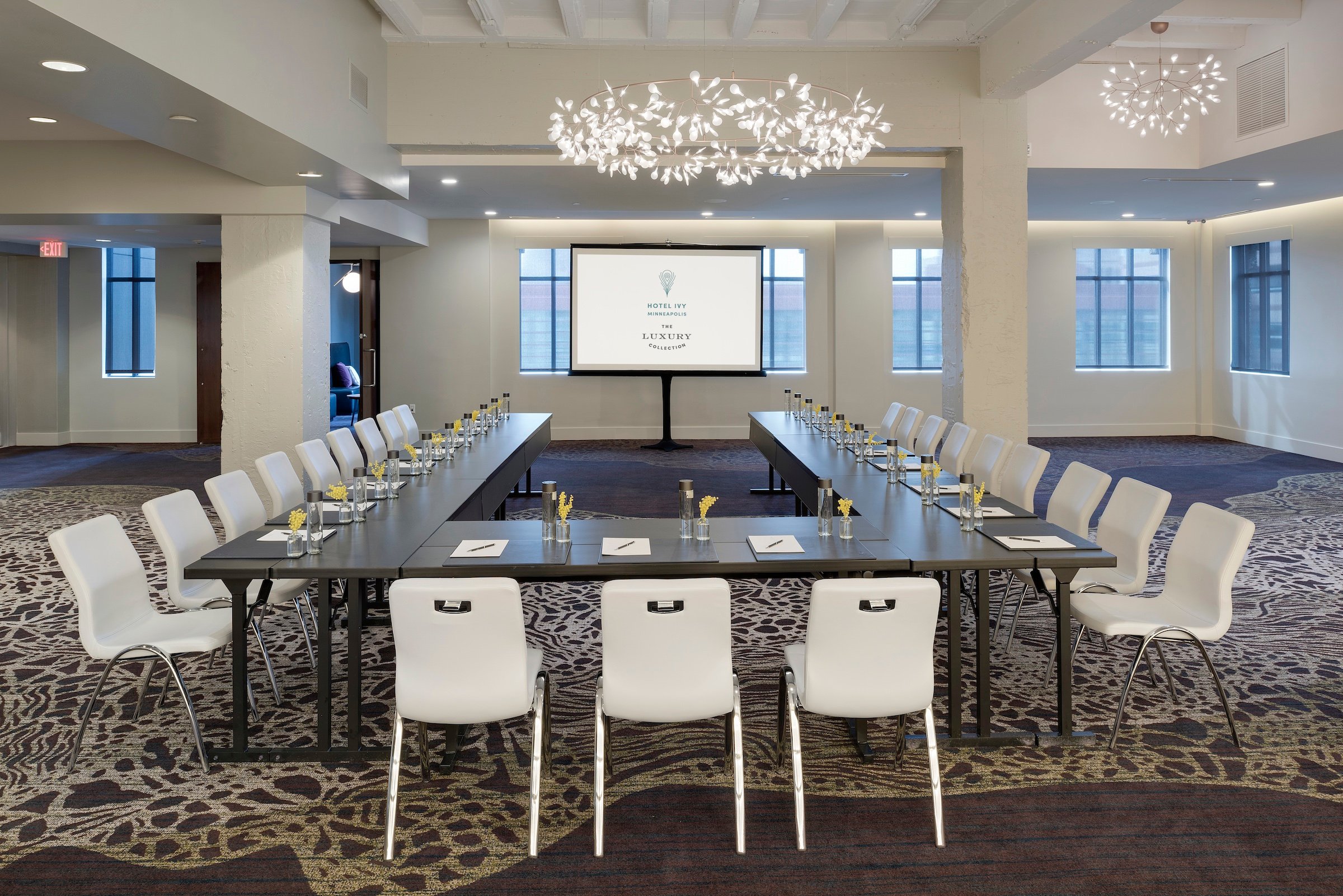 A spacious and elegantly designed meeting room at Hotel Ivy’s Grand Studio, set up in a U-shape conference style with white chairs, dark tables, and a presentation screen displaying the hotel's logo.