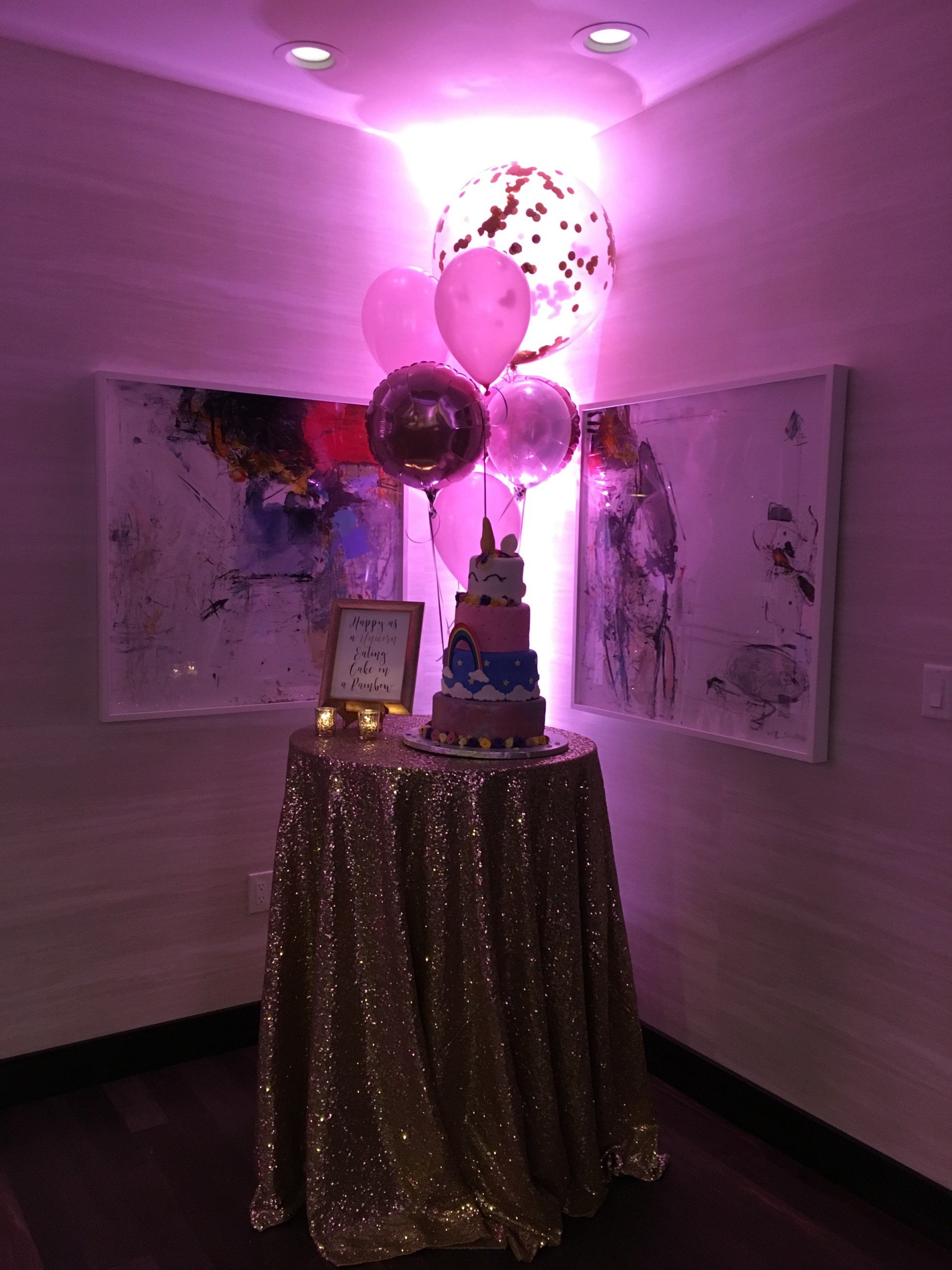 four layers of cake with characters for celebrations on the high table with shiny clothes and two paintings on the wall under pink lighting at Penthouse Suite of Hotel Ivy