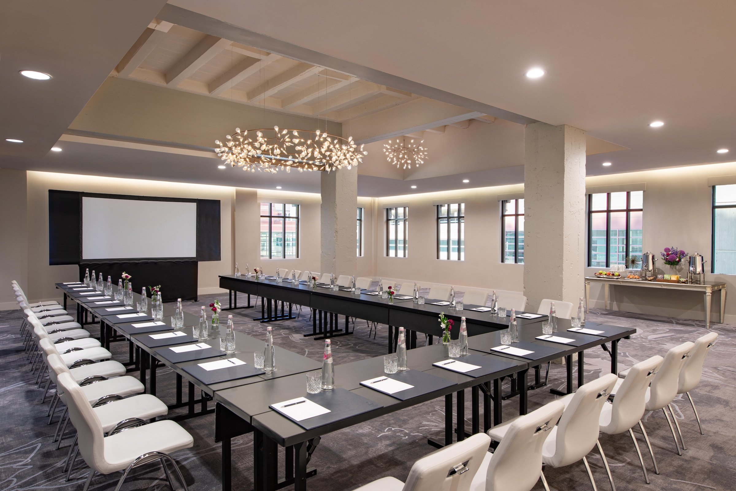 A U-shaped black desks and white chairs under unique lighting with a large monitor in a bright meeting venue