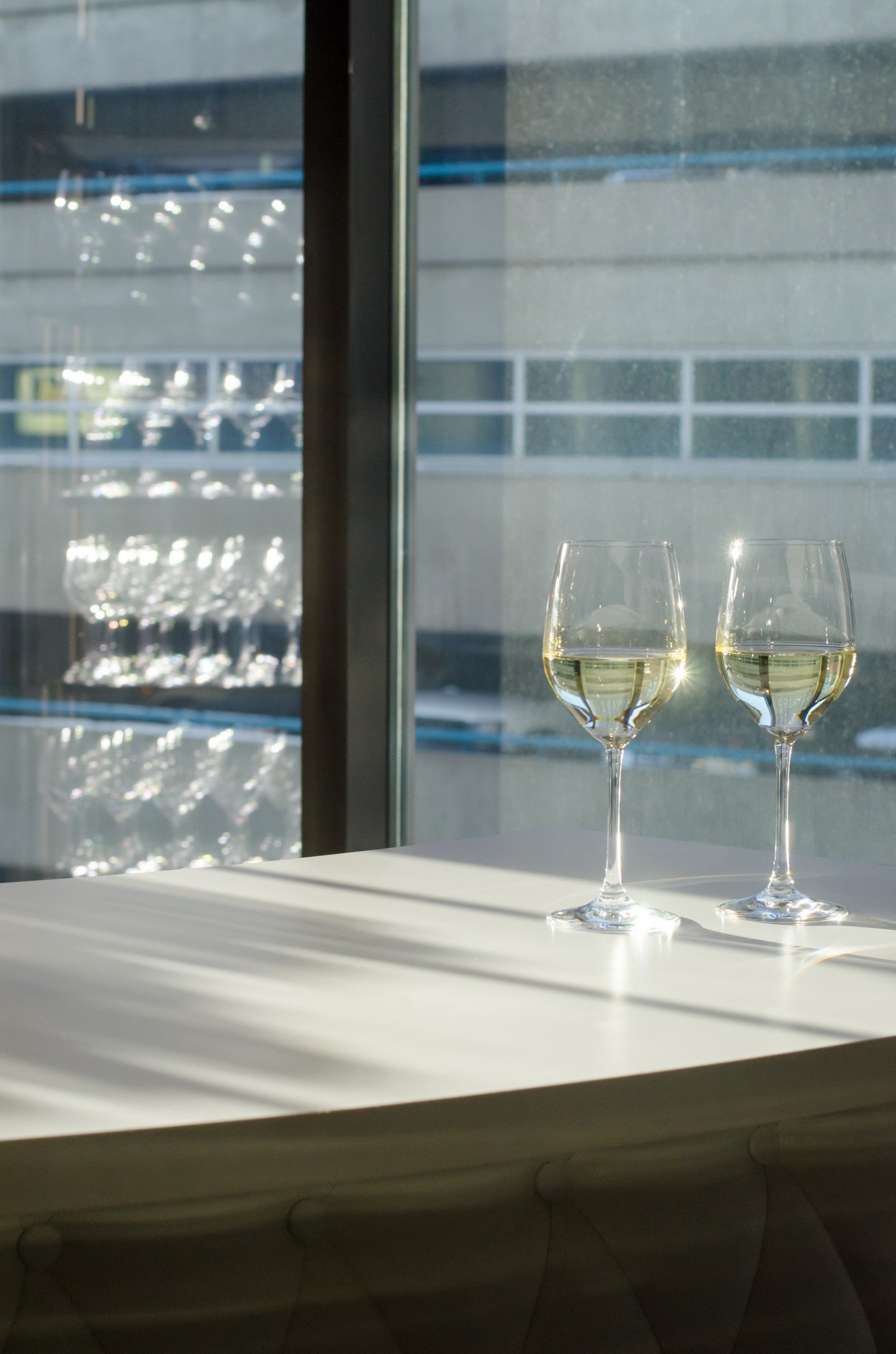 Two transparent glasses of white wine on a white table next to window, reflecting wine glasses selection