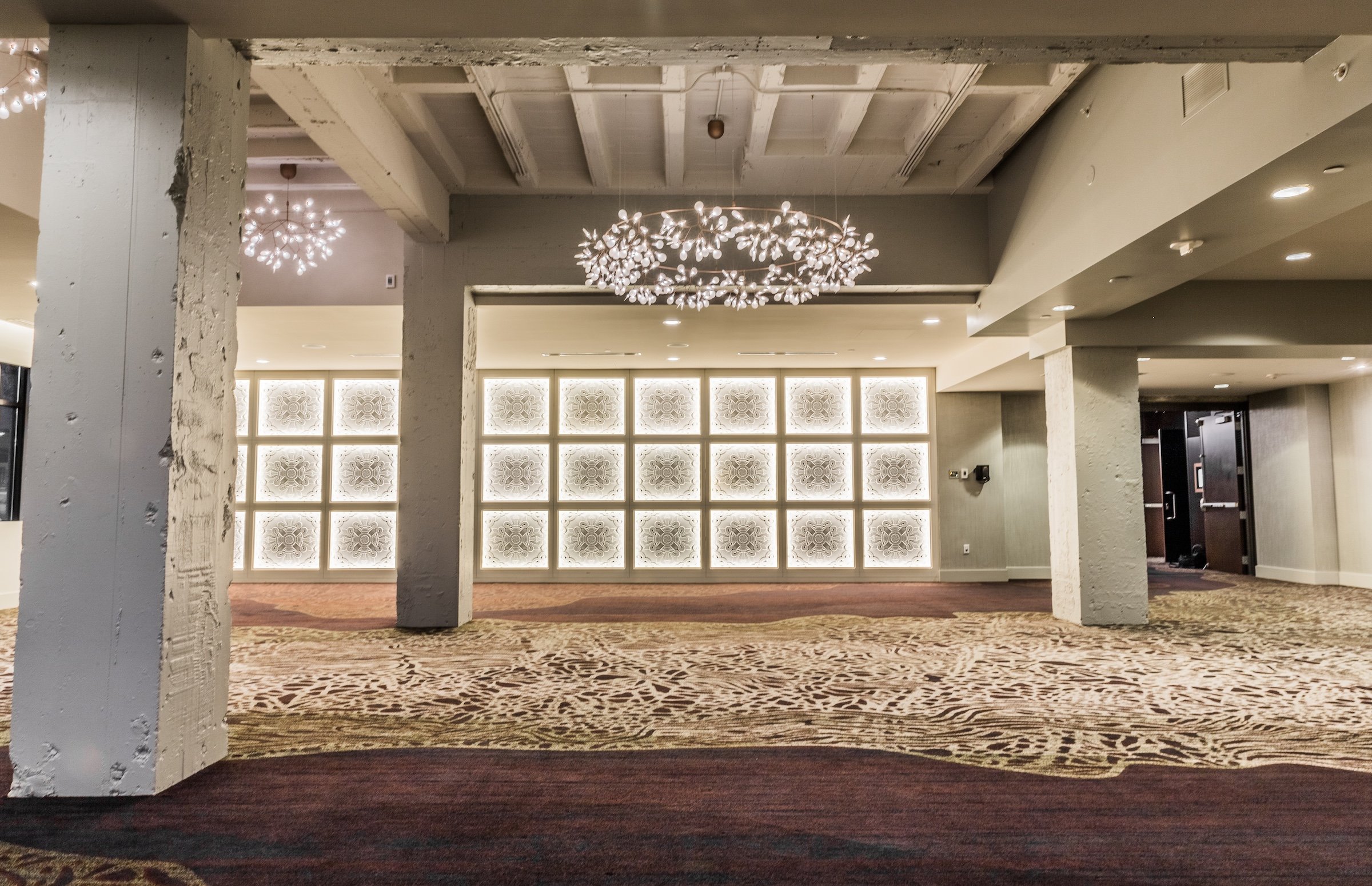 A well-lit event space in Hotel Ivy’s Grand Studio, featuring intricate chandeliers, high ceilings, and modern decor with neutral tones and artistic carpeting.