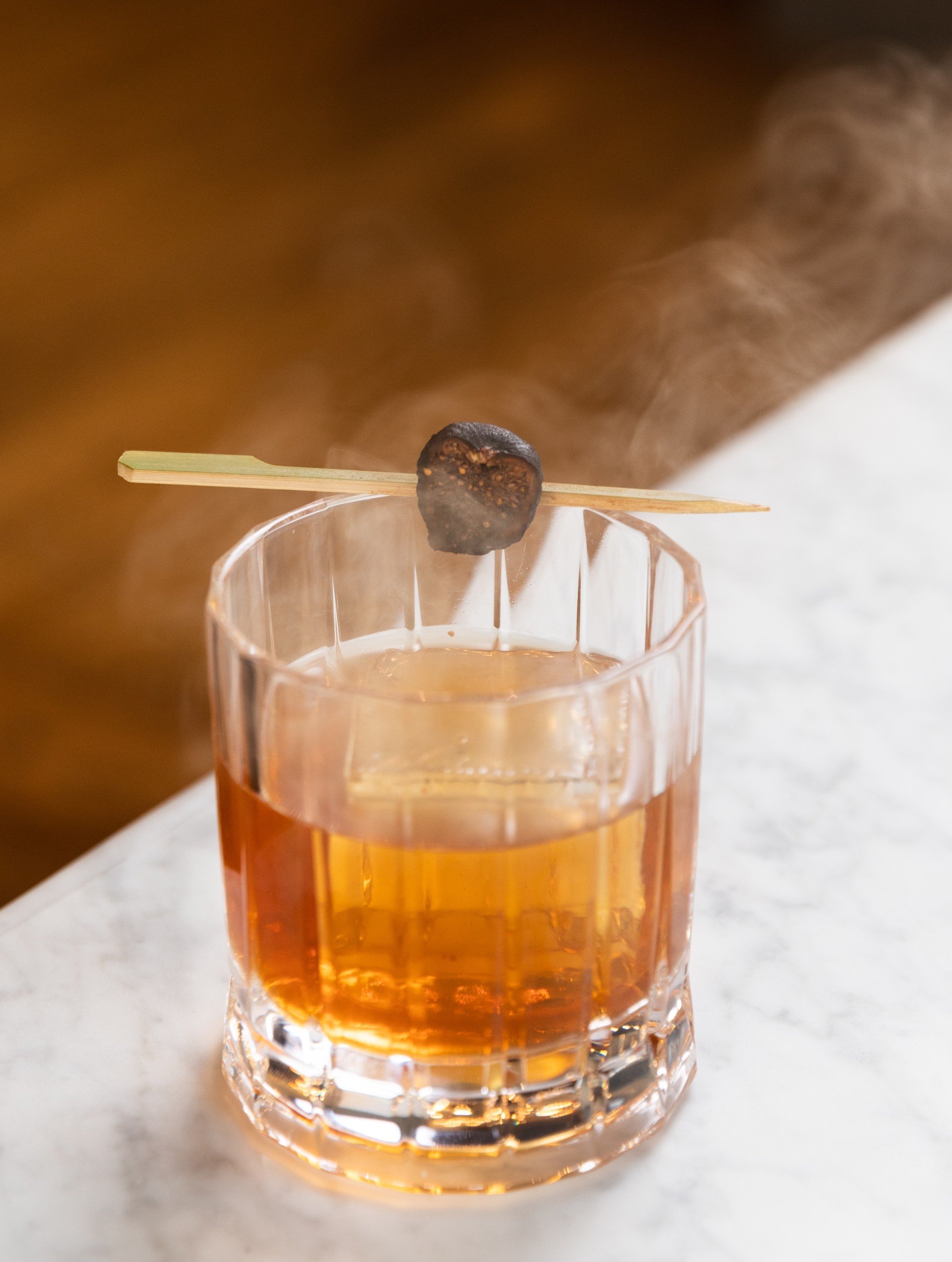 Smoked Fig Old Fashioned cocktail with a fruit on top on a white marble table at Breva Bar & Grill from Hotel Ivy