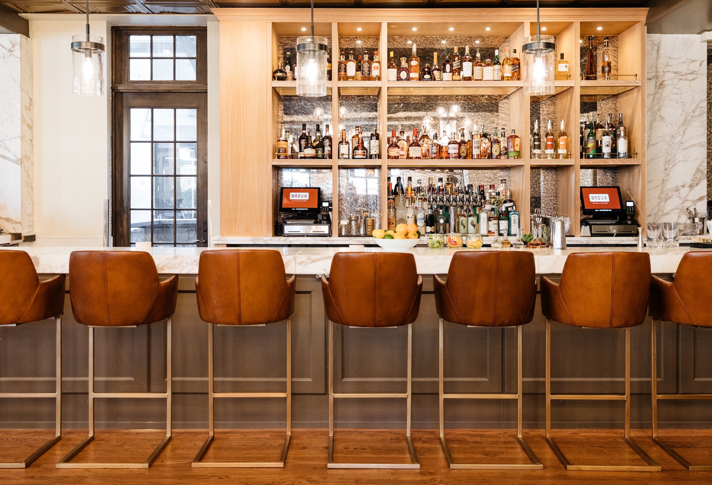 A bright bar area with white bar table and leather chairs displaying various liquors at Breva