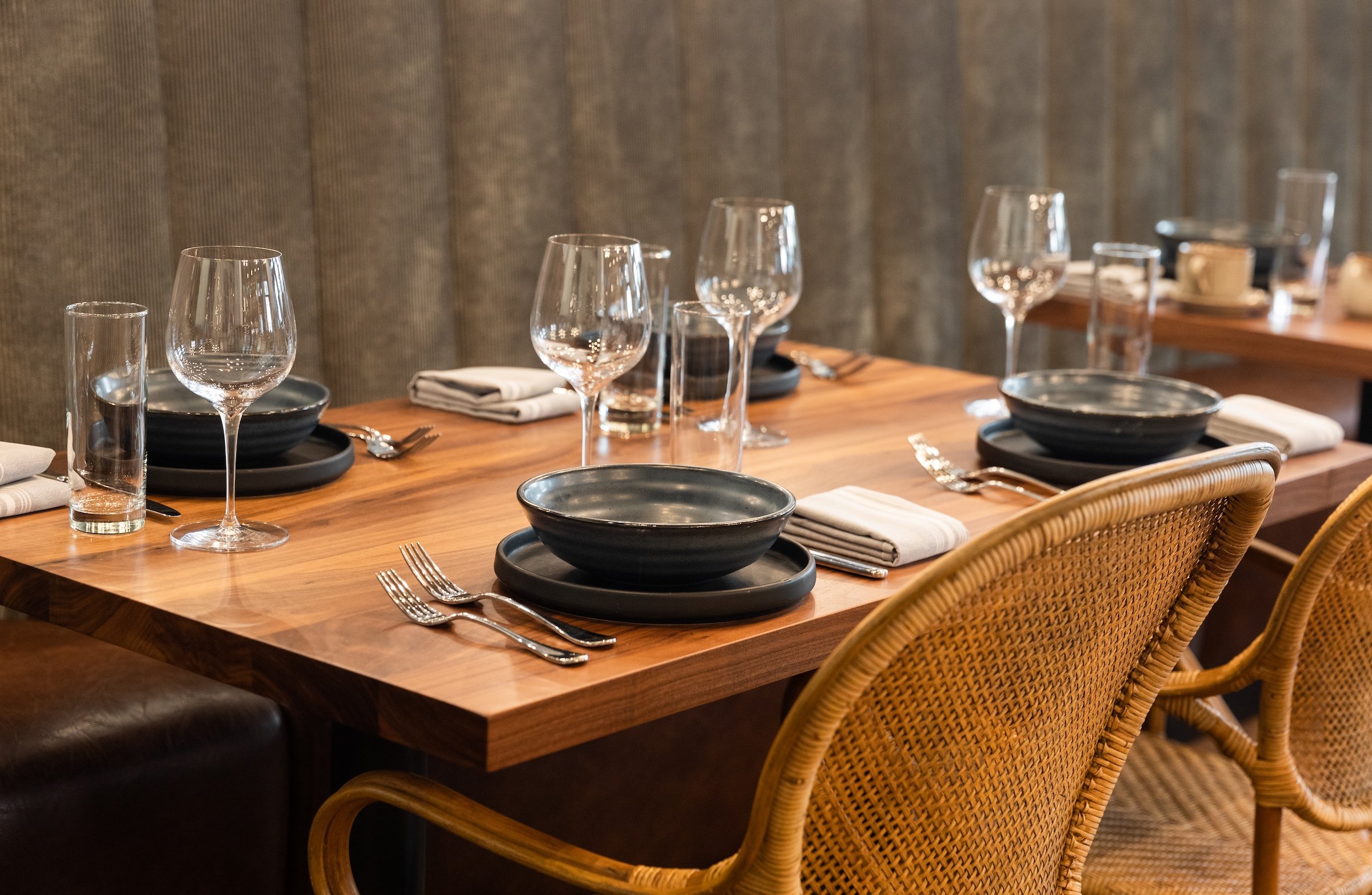 Wooden tables with table settings, ceramic bowls and plates, warm toned napkins, transparent water glass and wine glass on wooden tables with rattan chairs at Breva Bar & Grill from Hotel Ivy