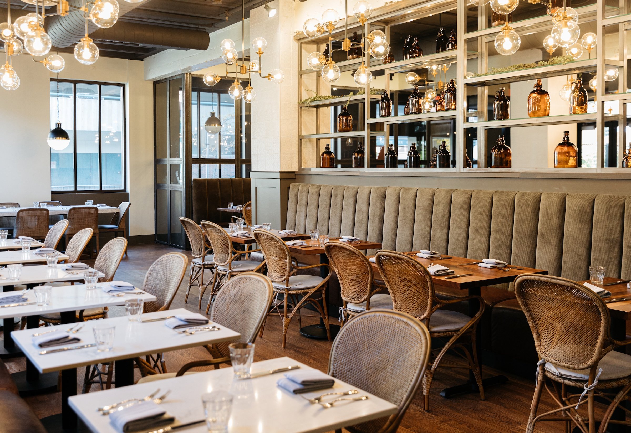 Wooden dining tables, white marble tables, brown rattan chairs, and brown leather couch next to a bottles decoration under round stylish lightbulbs at Breva Bar & grill from Hotel Ivy