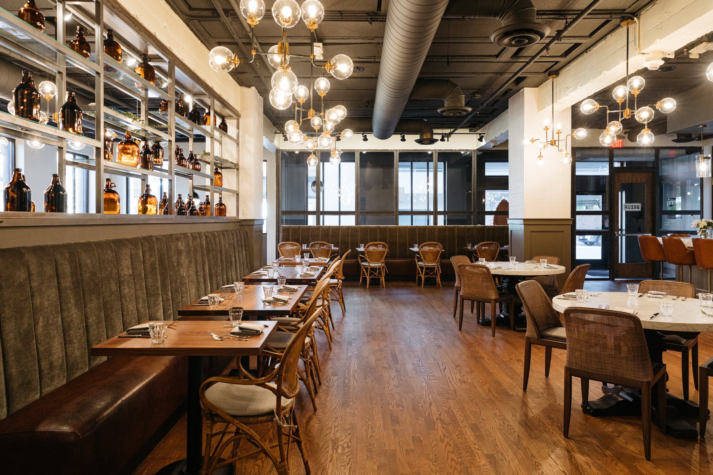 A modern stylish interior of Breva, wooden tables with tidy settings under round lightbulbs with pipe in a bright dining area at Hotel Ivy