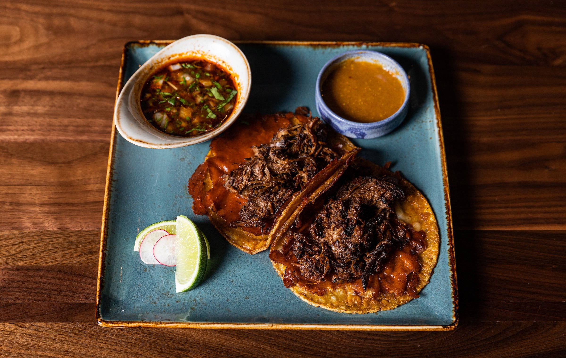 Beef Birria tacos with salsa sauce and limes from a stylish blue square plate on a wooden table at Masa & Agave of Hotel Ivy