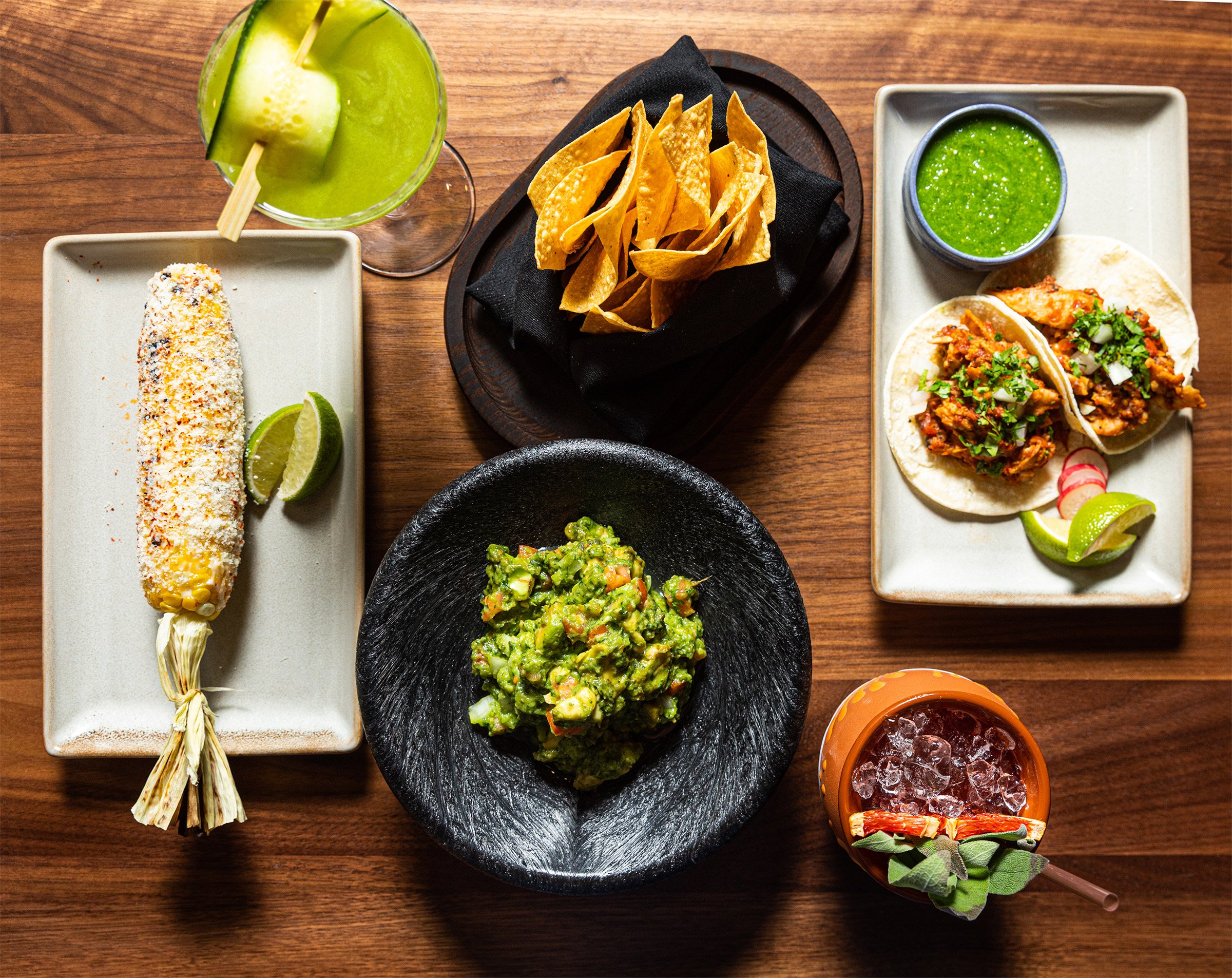 A beautifully plated Mexican feast on a wooden table, featuring elote guacamole in a black stone bowl, crispy tortilla chips, and two tacos filled with seasoned meat, topped with cilantro and onions at Brave Bar & Grill from Hotel Ivy