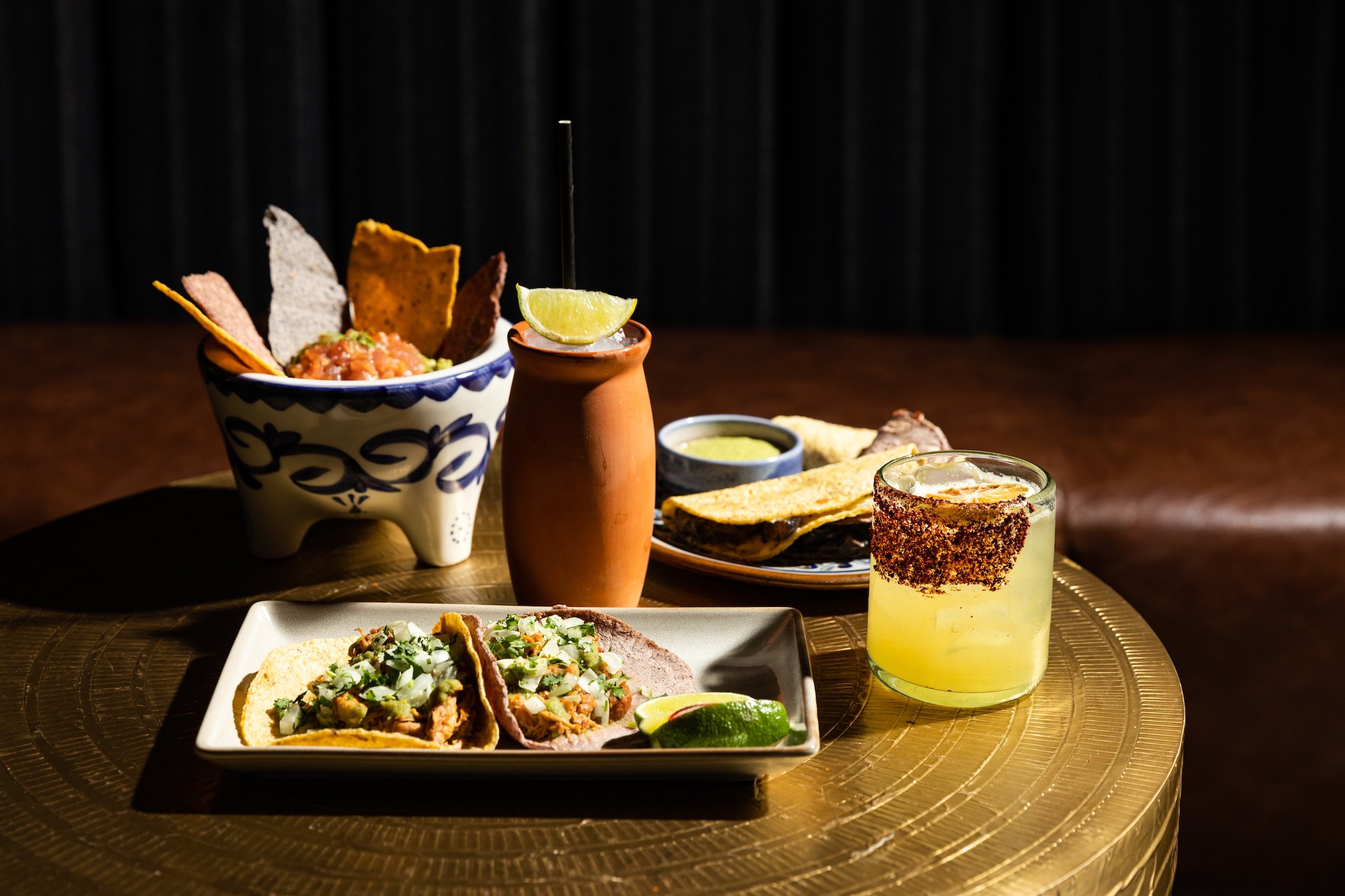 Colourful dishes of tacos, mexican bowl, nachos with cocktails in unique cups on a golden table next to brown leather couchat Masa & Agave from Hotel Ivy