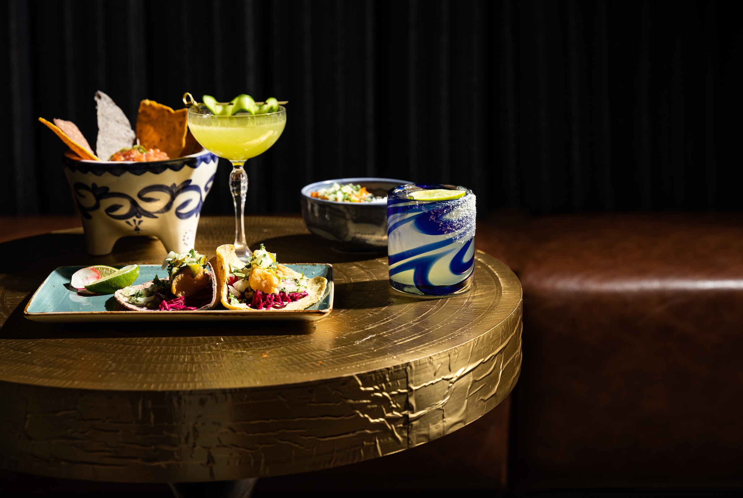 Colourful dishes of tacos, mexican bowl, nachos, cocktails on a golden table next to brown leather couchat Masa & Agave from Hotel Ivy