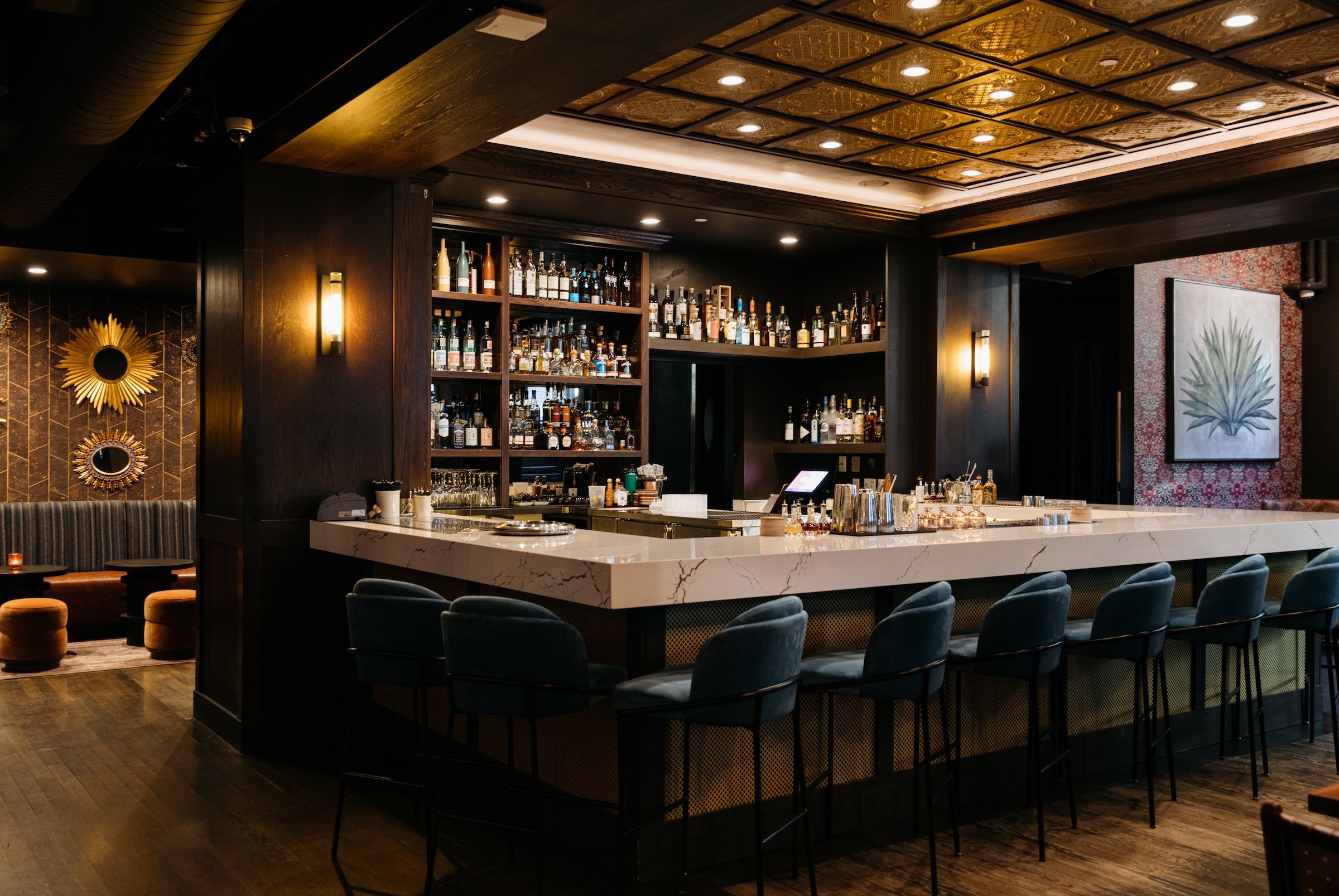 A decent selection of liquor and a marble white bar table with light blue high chairs under darken warm lighting next to Agave drawing and round mirrors representing sun at Masa & Agave from Hotel Ivy