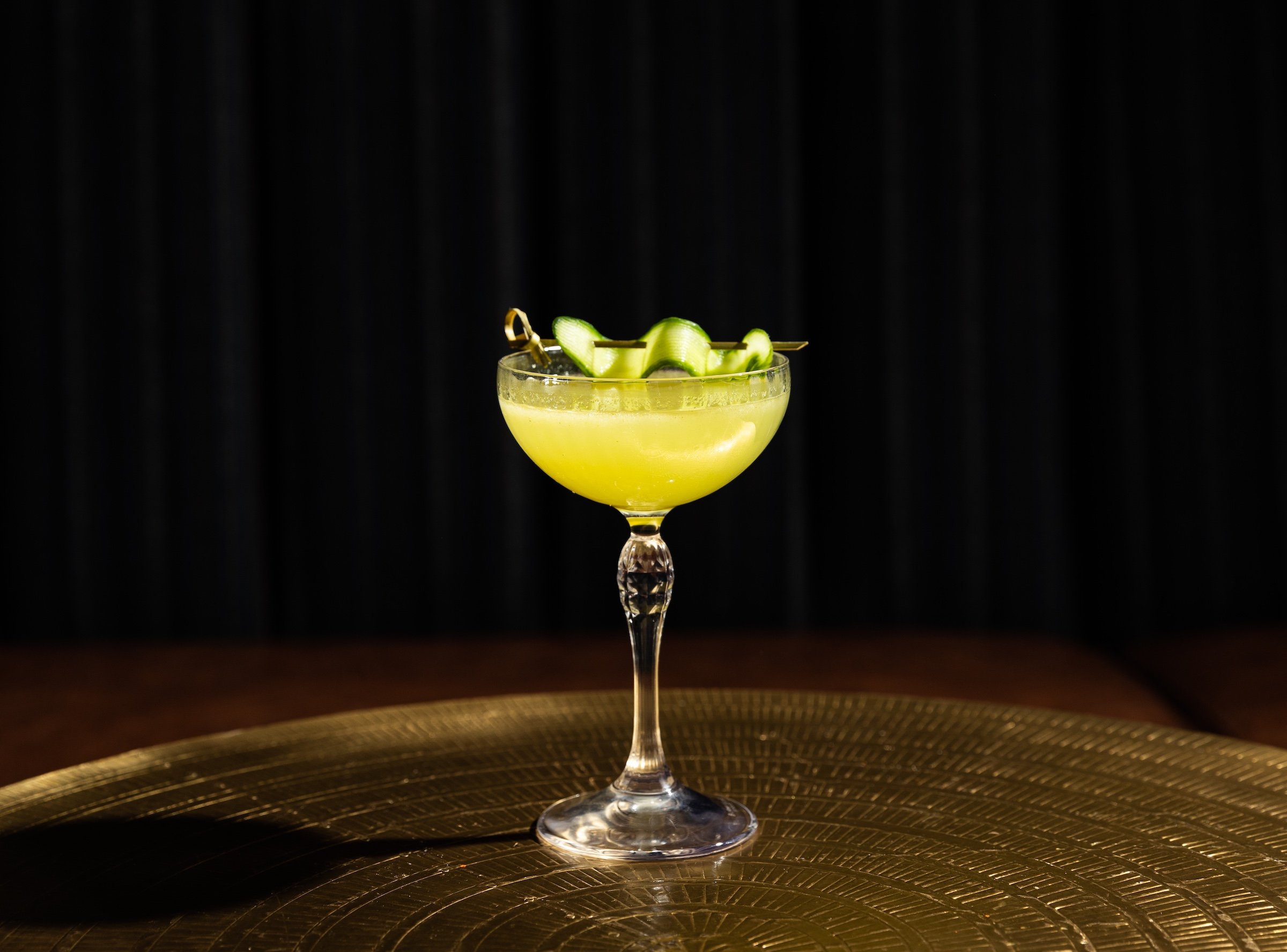 A green cocktail decorated with cucumber in a elegant wine glass on a golden table in a dark room at Masa & Agave from Hotel Ivy