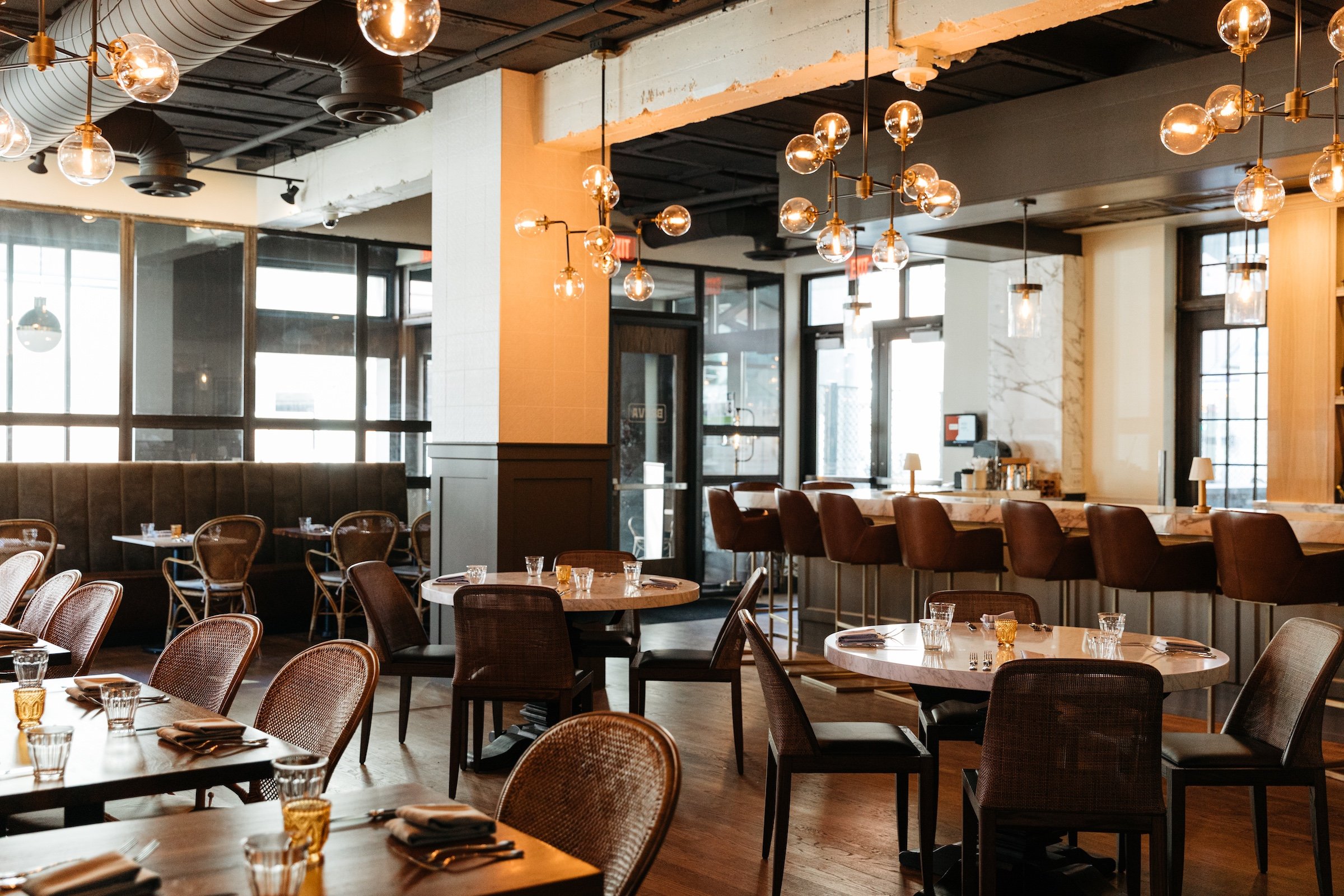 A marble bar table with brown leather high chairs and dining tables with rattan chairs next to large glass entrance in a bright space, Breva Bar & Gill from Hotel Ivy